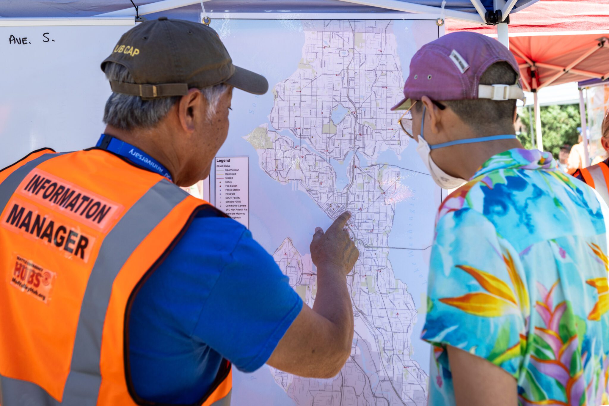 The back of two people looking at a posted map. One person is wearing a bright orange safety vest that says "Information Manage" 