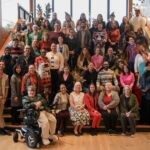 A large group of people posing on a large, open, wooden stairwell in a brightly lit room