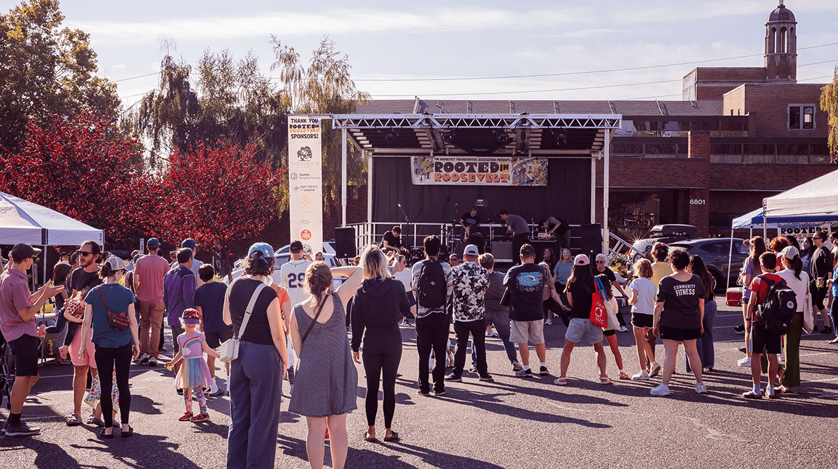 Groups of people gathered in front of an outdoor stage on a sunny day at a neighborhood block party.