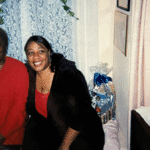 A vintage photo - circa 1990s of two Black women sitting in chairs smiling with blue lace curtains in the background