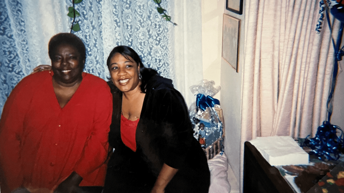 A vintage photo - circa 1990s of two Black women sitting in chairs smiling with blue lace curtains in the background