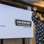 A Black woman wearing a black dress with white polka dots passionately singing into a microphone with one hand raised in the air. A projector screen appears in the background with a logo with text "Black History Month. Rooted, Resilient, and Rising. Lift Every Voice & Sing. Josephine Howell."