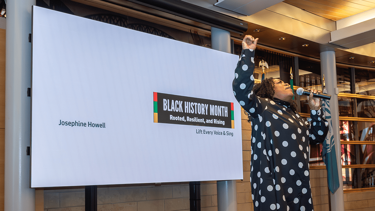 A Black woman wearing a black dress with white polka dots passionately singing into a microphone with one hand raised in the air. A projector screen appears in the background with a logo with text 