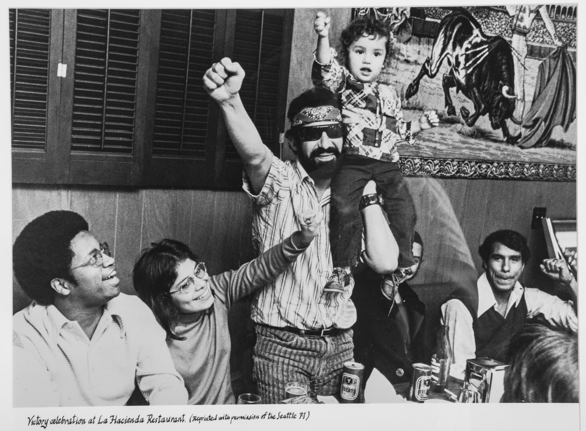 Black and white photo of man standing with sunglasses holding a child on his shoulder and raising a fist in the air while people seated next to them look up smiling