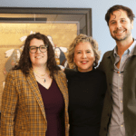 A woman with long, curly brown hair and glasses, a woman with short light hair, and a tall man with short brown hair stand together in an office.