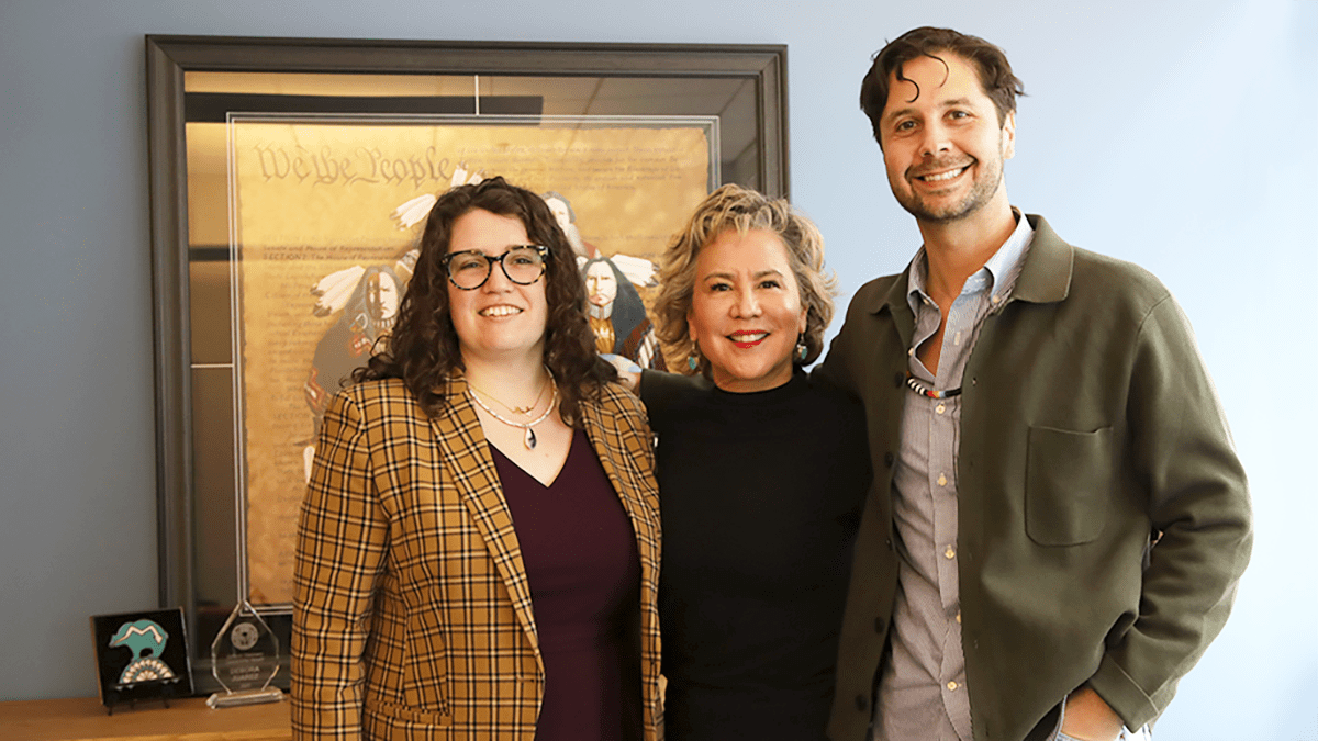 A woman with long, curly brown hair and glasses, a woman with short light hair, and a tall man with short brown hair stand together in an office.