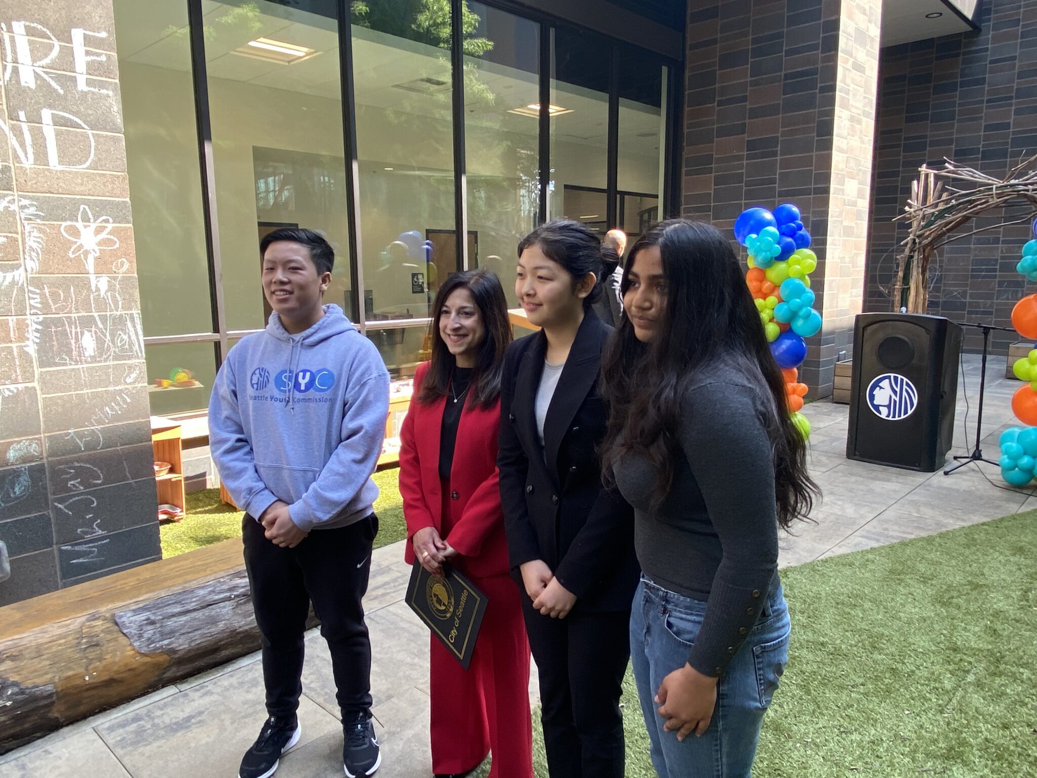 Three youth standing with a woman with medium length black hair, wearing a red pant suit stand together on an patio
