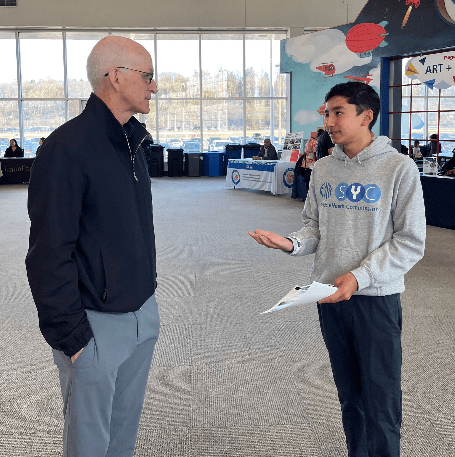 An older man with gray hair and glasses wearing a black sweater and slacks speaks to a youth with brown skin and short black hair wearing a gray sweatshirt that says "Seattle Youth Commission"
