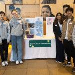 Six teens stand next to a table that has a presentation board atop and a sign that reads "Seattle Youth Commission"