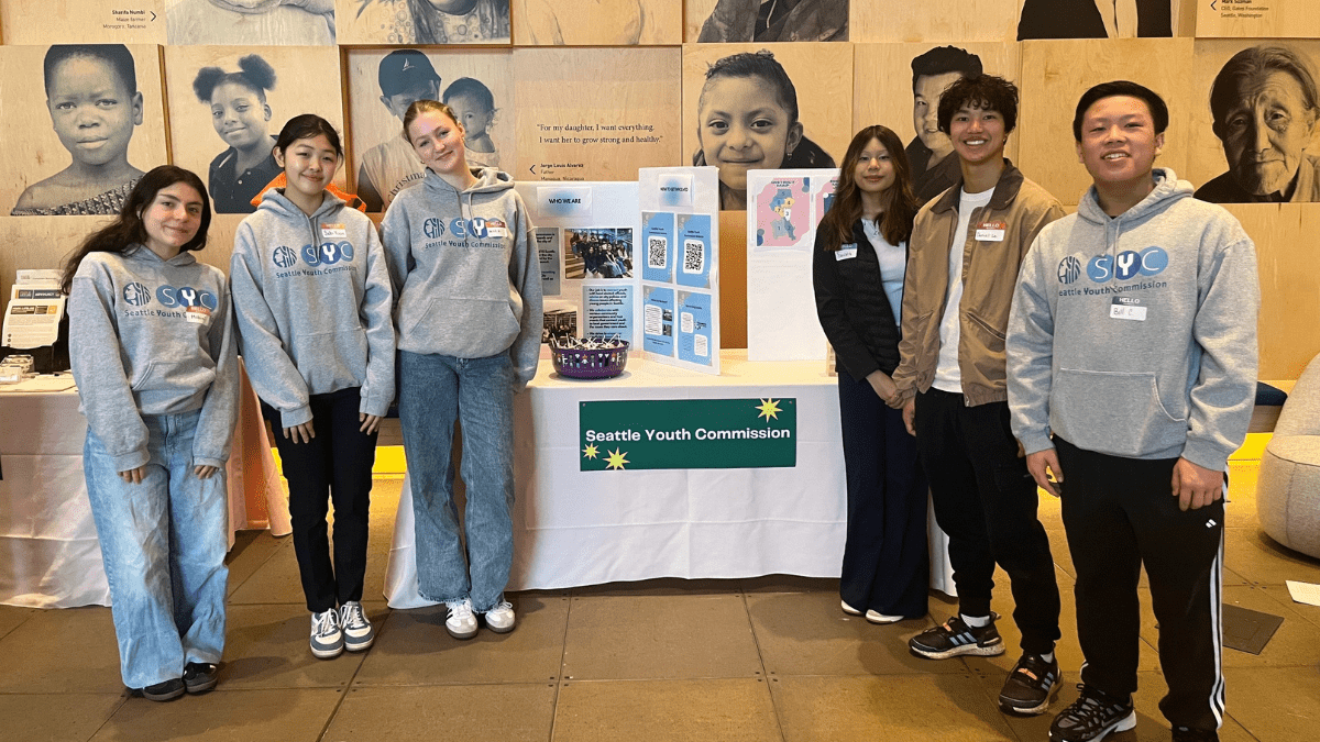Six teens stand next to a table that has a presentation board atop and a sign that reads