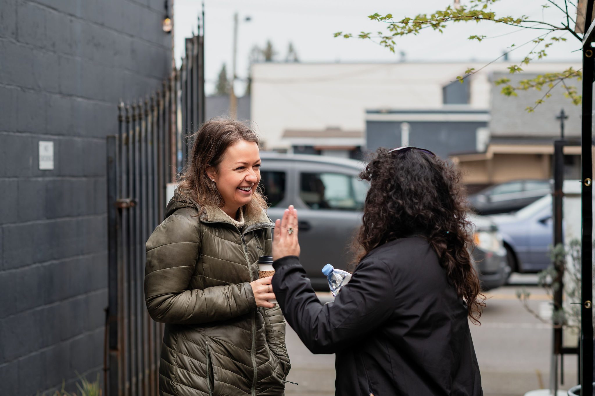 A person with a brown puffer coat and medium length brown hair standing outside talking to a person with long wavy black hair 