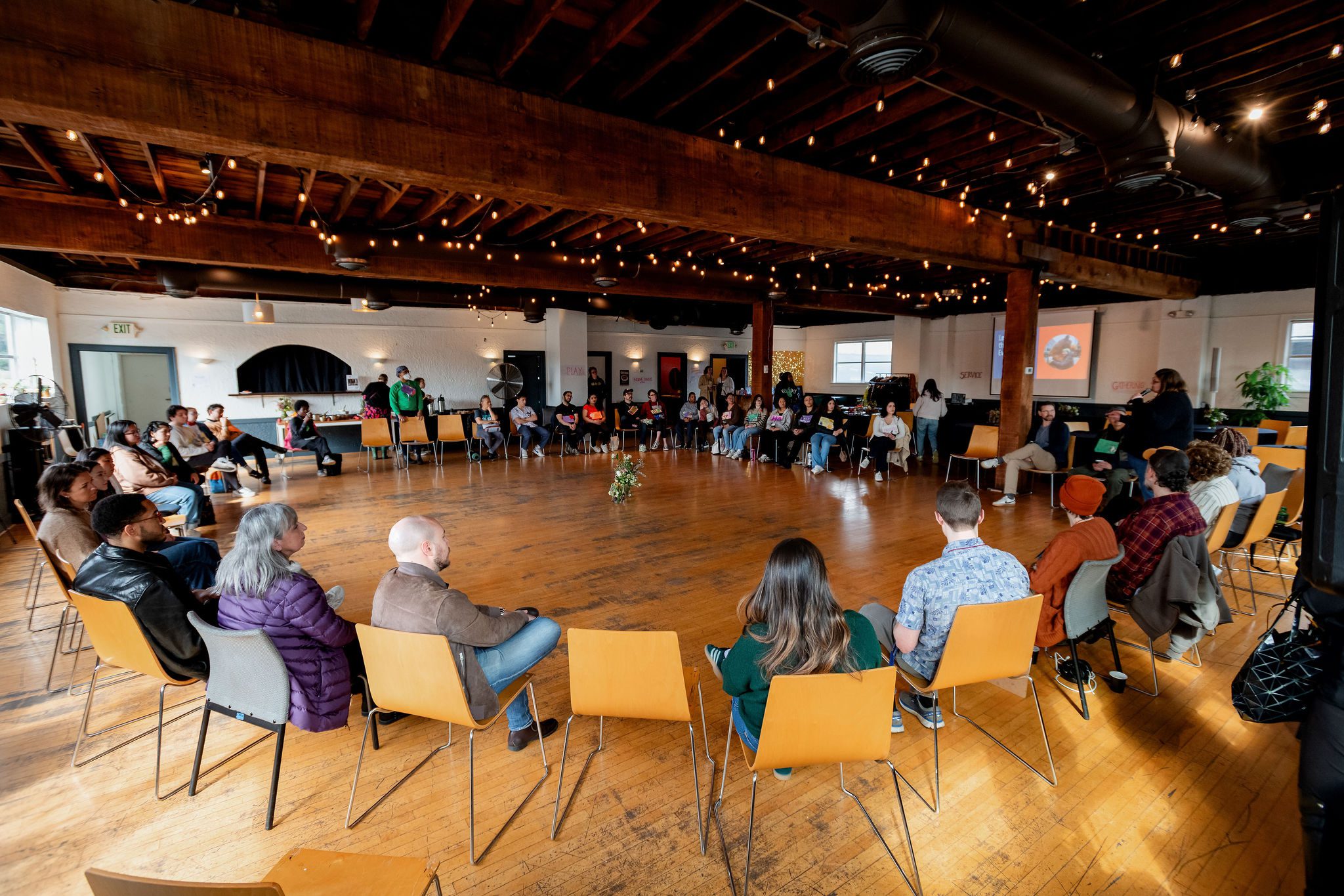 A large group of people sitting in chairs in a circle in a large venue with wood floors