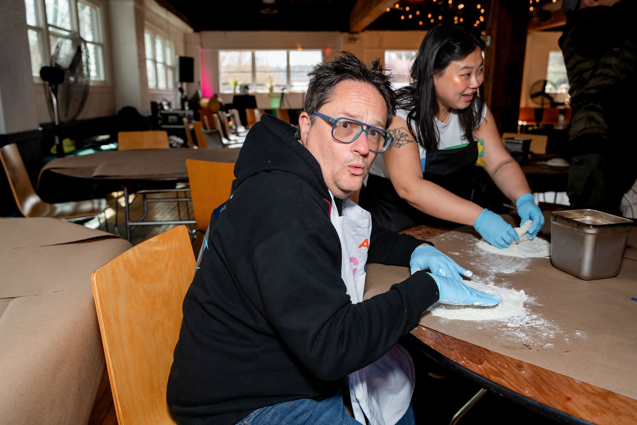 A person with short black hair and glasses sitting at a table kneading pizza dough