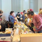 A group of people standing along both sides of a long table holding plant starts and looking at packs of seeds laid out on the table.