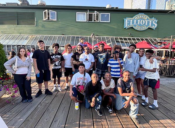 A large group of youth pose together on a dock in front of a building with green siding and a sign reading "Elliott's Oyster House" 