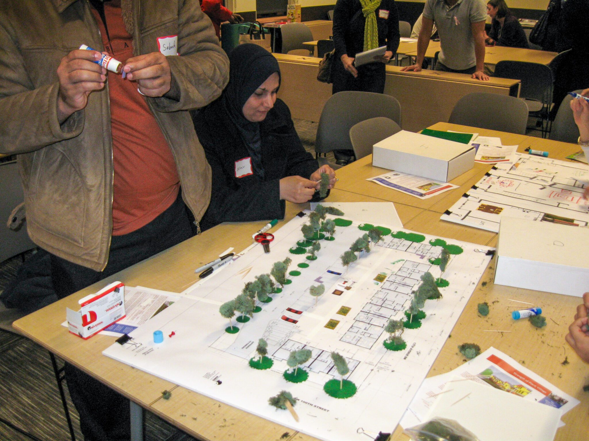 Two people sitting at a table places small crafted trees around a blueprint of buildings