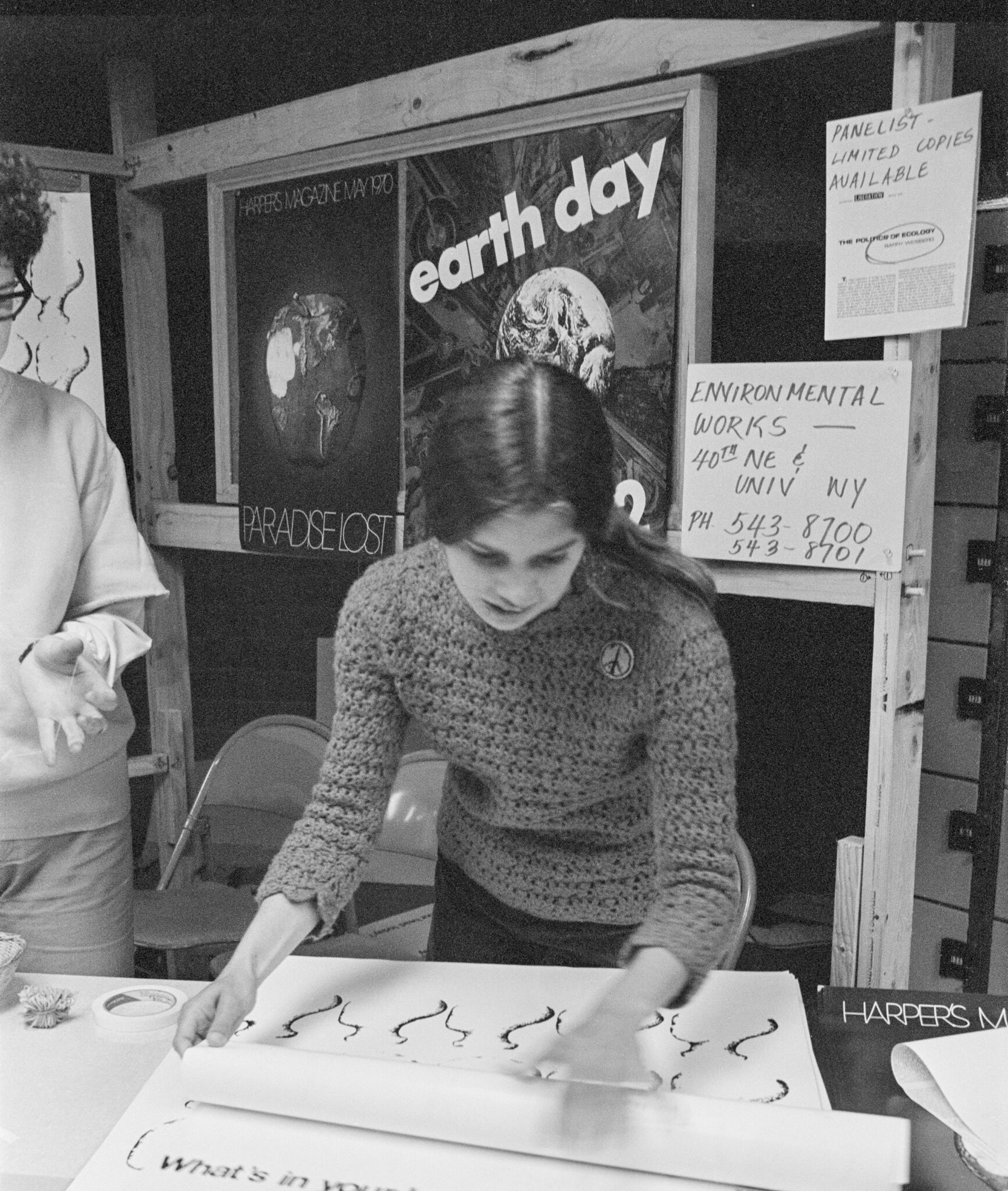 Black and white photo of a young woman looking at posters on a table with a sign behind her that says "Earth Day"