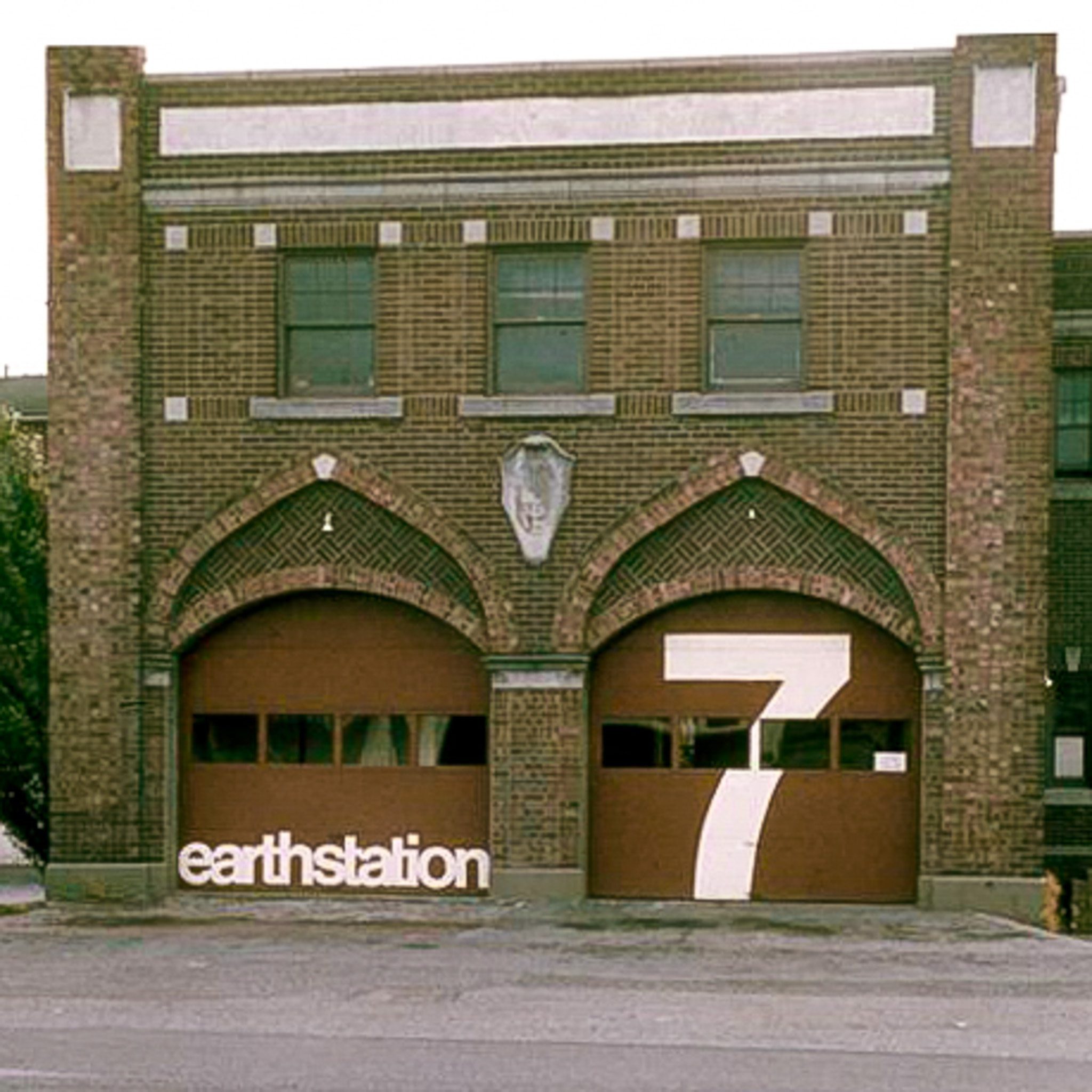 Vintage, sepia toned photo of a brick, two-story building with two large garage doors in front the "Earthstation 7" painted in white across them