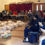 People sitting in chairs around boxes of food lined up in the center of a large room