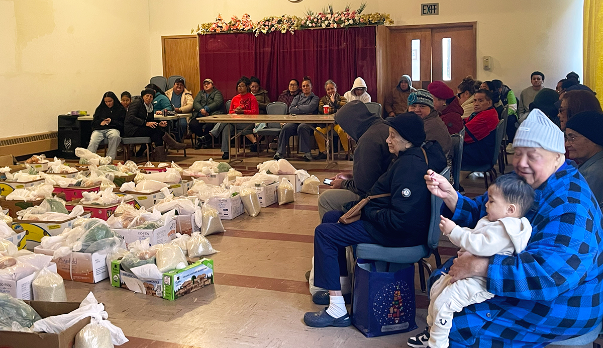 People sitting in chairs around boxes of food lined up in the center of a large room