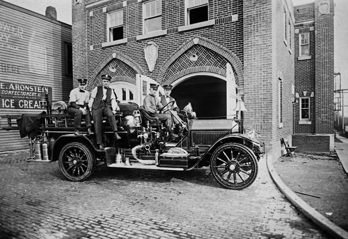 A black and white photo from the early 1900's with five people sitting in a vintage fire truck in front of a brick firehouse