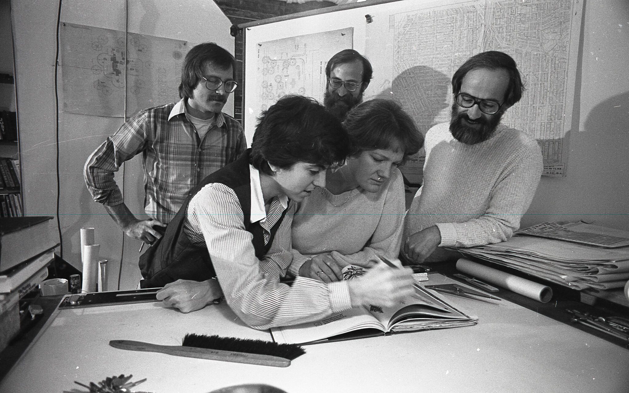 Black and white photo of five people around a desk looking down at a book with blueprints and maps hanging on the walls around them