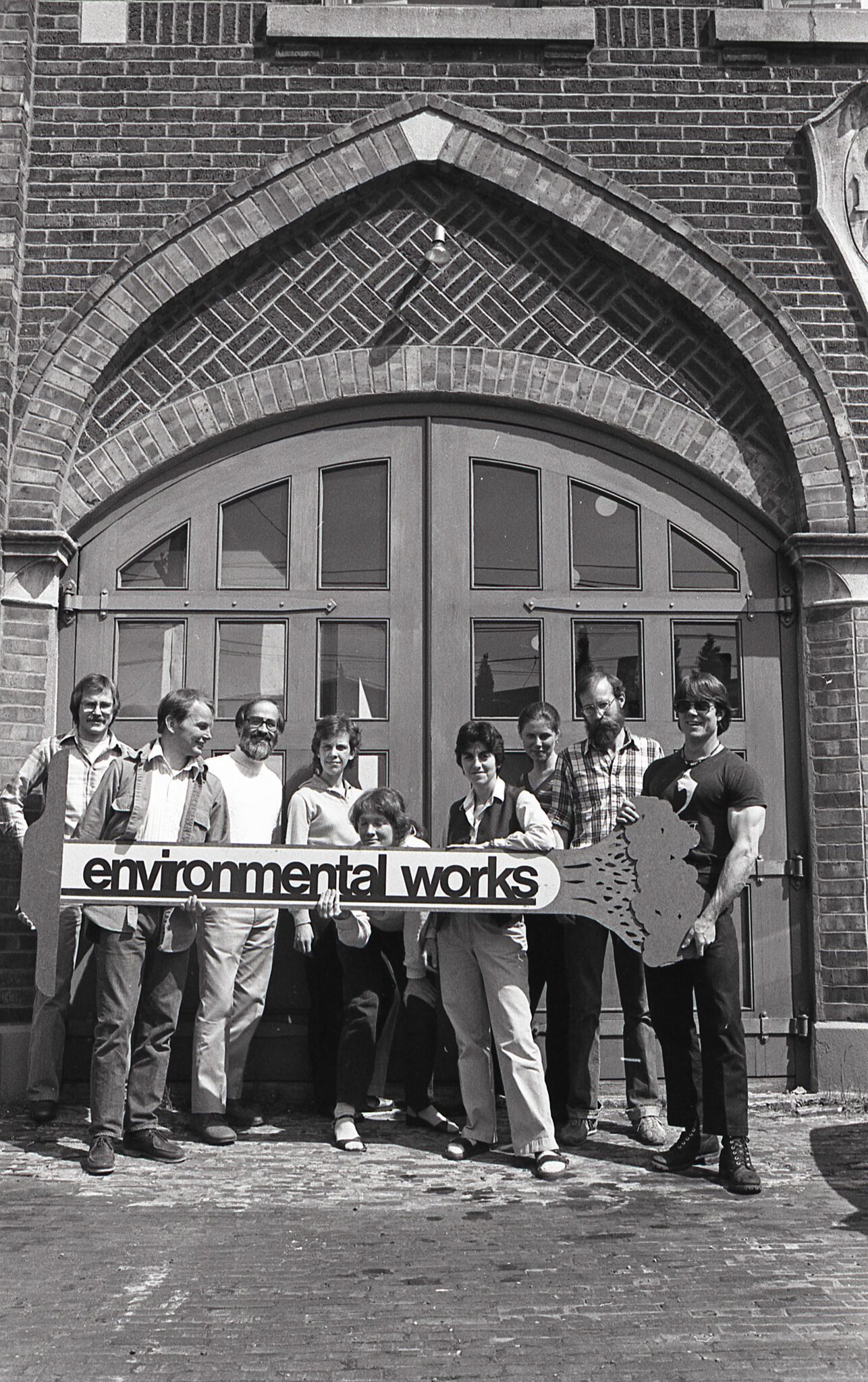 Black and white photo of a group of people standing in front of brick building with a large bay door holding a sign that says "Environmental Works"