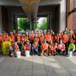 A large group of people wearing bright orange vests with plastic yellow garbage bags, buckets, and trash grabbers