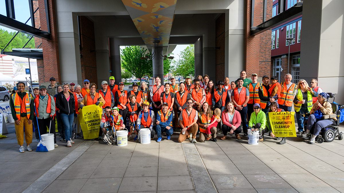 A large group of people wearing bright orange vests with plastic yellow garbage bags, buckets, and trash grabbers
