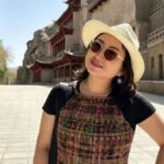 A Chinese woman wearing a brimmed straw hat and sunglasses smiling and standing on a brick walkway with Chinese architectural buildings in the background
