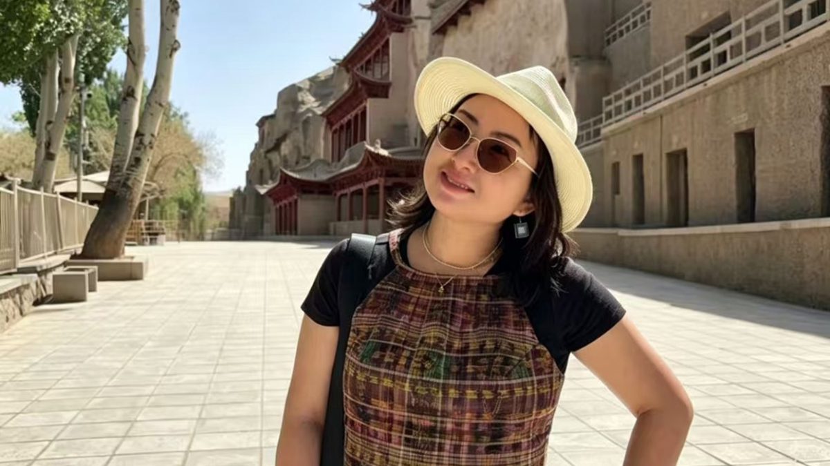 A Chinese woman wearing a brimmed straw hat and sunglasses smiling and standing on a brick walkway with Chinese architectural buildings in the background