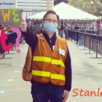 A Chinese man wearing a orange safety vest and blue medical mask stands outside a stadium with his arm extended giving a thumbs up