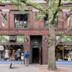 Exterior of a two story brick building with a person standing on the sidewalk looking into the large windows of a retail space on the ground level