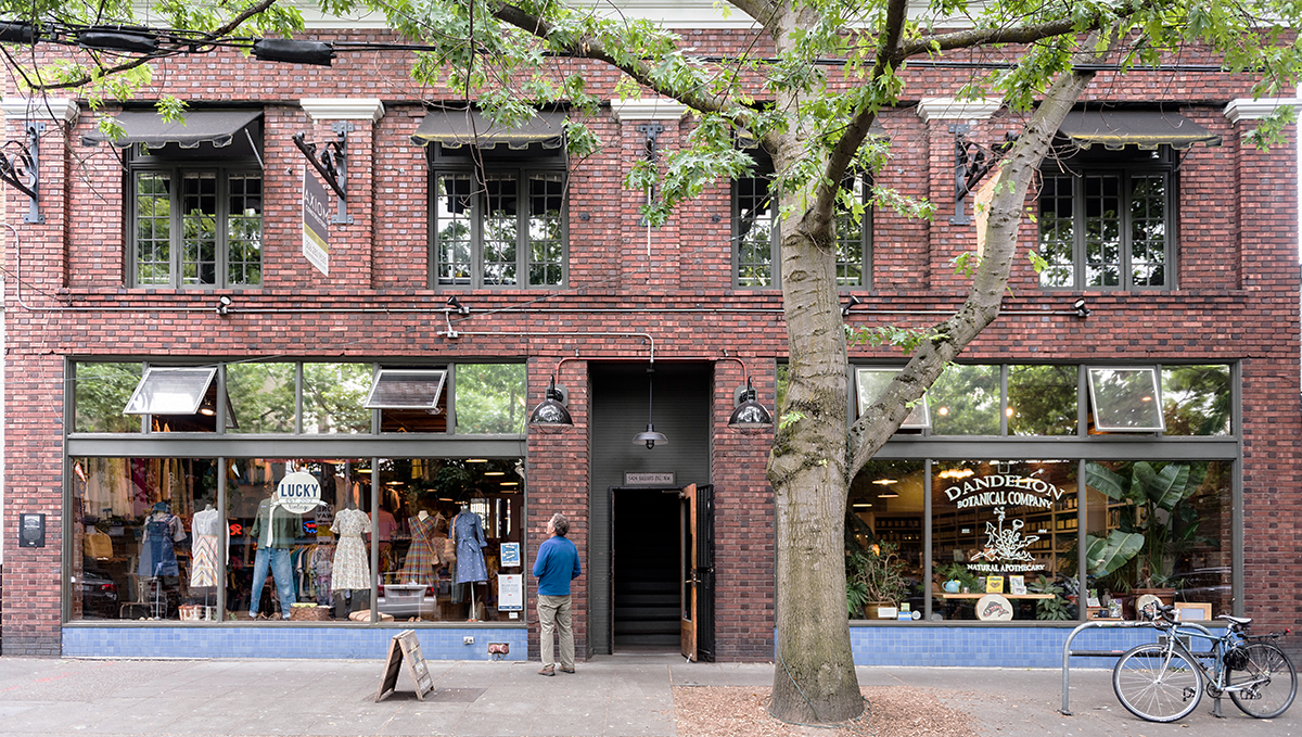 Exterior of a two story brick building with a person standing on the sidewalk looking into the large windows of a retail space on the ground level