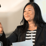 A Vietnamese woman with long black hair wearing a black striped shirt and black suit jacket smiling and reaching for a microphone