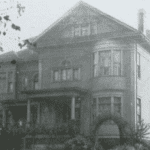 vintage, black and white photo of a wood frame house composed in a neo-classical manner. Its two story height is capped by a deeply recessed classic pediment. A rounded bay window rises two stories, ending under the cornice.