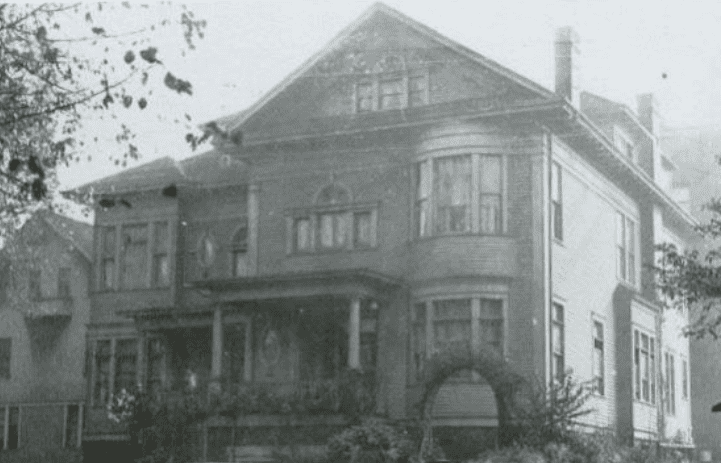 vintage, black and white photo of a wood frame house composed in a neo-classical manner. Its two story height is capped by a deeply recessed classic pediment. A rounded bay window rises two stories, ending under the cornice.