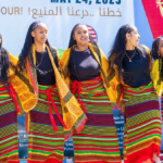 Six people wearing traditional African skirts, black shirts, and yellow scarfs draped over their shoulders stand together on a stage