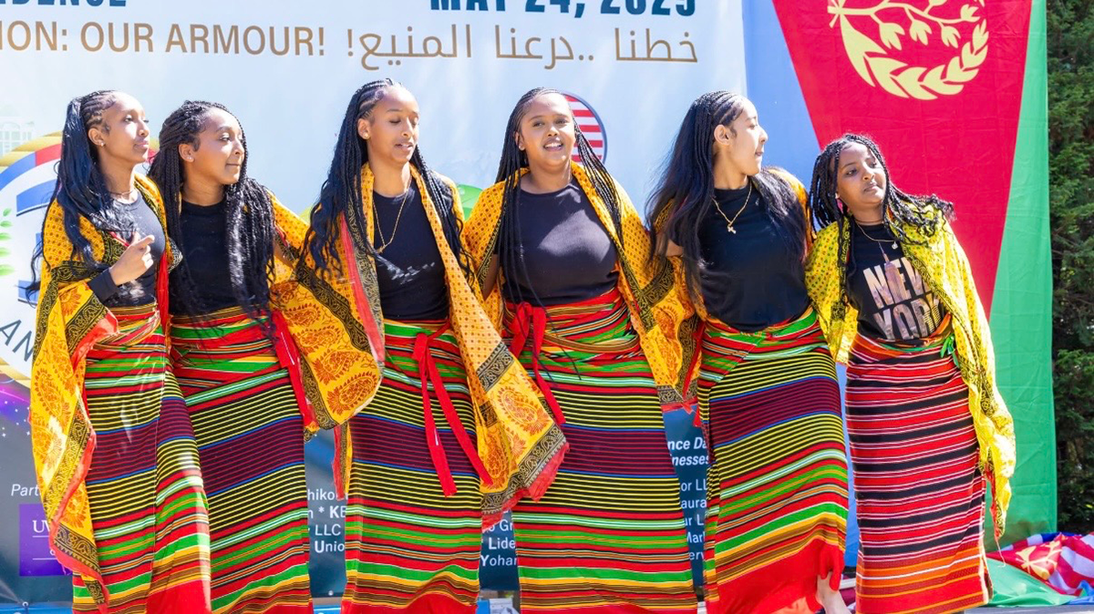 Six people wearing traditional African skirts, black shirts, and yellow scarfs draped over their shoulders stand together on a stage