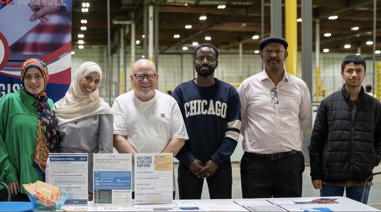 Six people of varying ages and ethnicities stand in a line behind a table with informational pamphlets