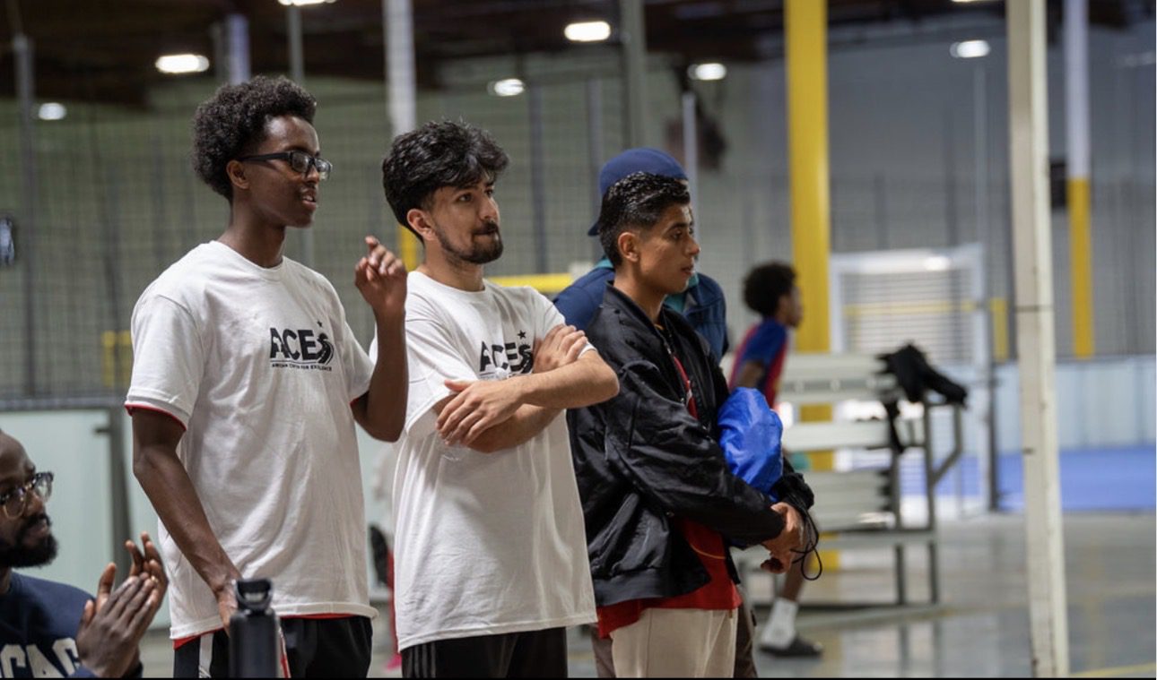 Three young men stand on the sideline of an indoor soccer field