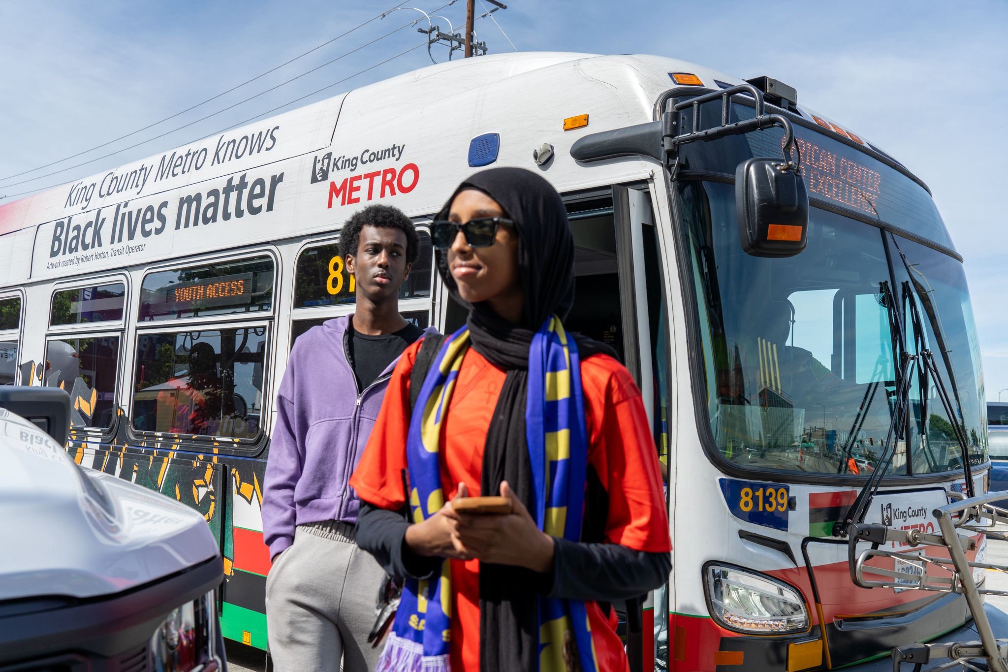 A young woman wearing a hijab, sunglasses, and a blue scarf stands next to a young Black man wearing a purple shirt just outside a city bus.