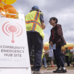 A white sandwich board sign reads "Community Emergency Hub Site" and a person in a high viz vest stands next to it speaking to another person.