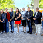A group of eleven people stand together on an outdoor patio with trees and blue sky visible in the background.