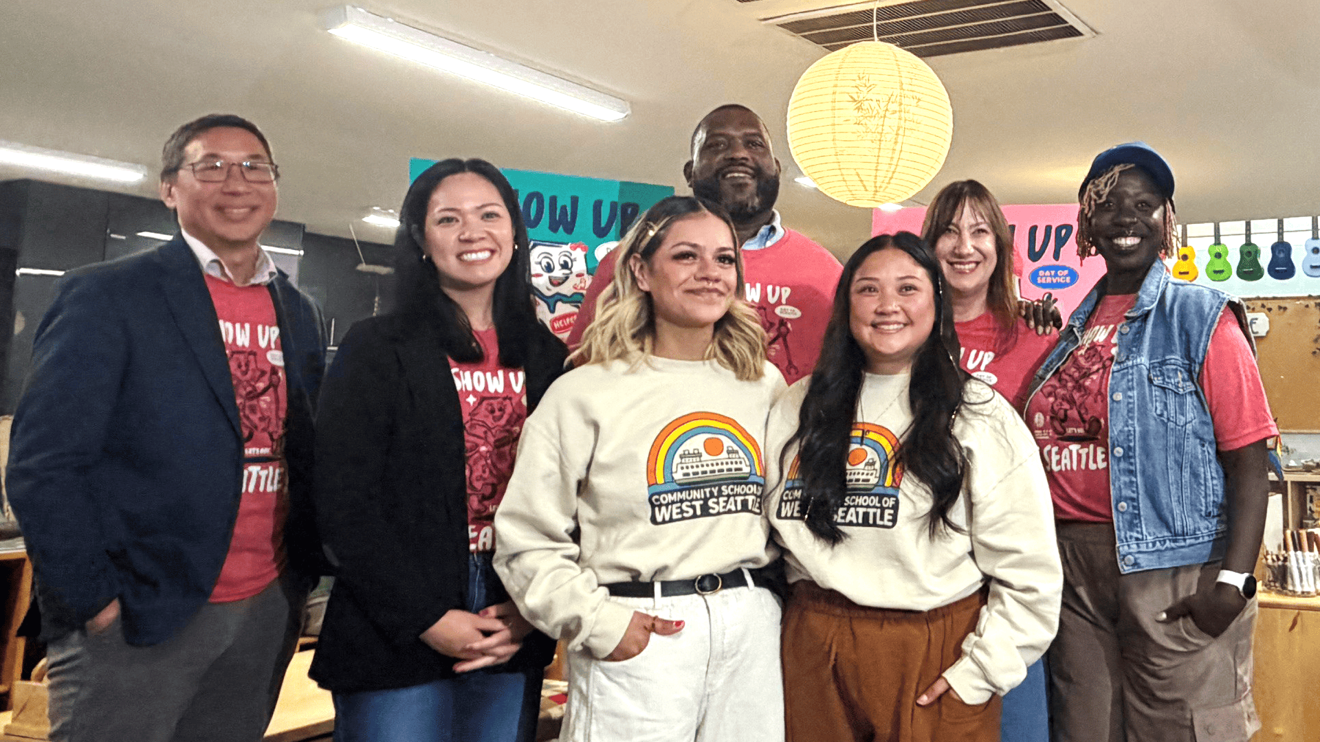 Seven people stand together posing for a photo. Five of them are wearing matching red shirts and the other two wearing white sweatshirts with a rainbow. 