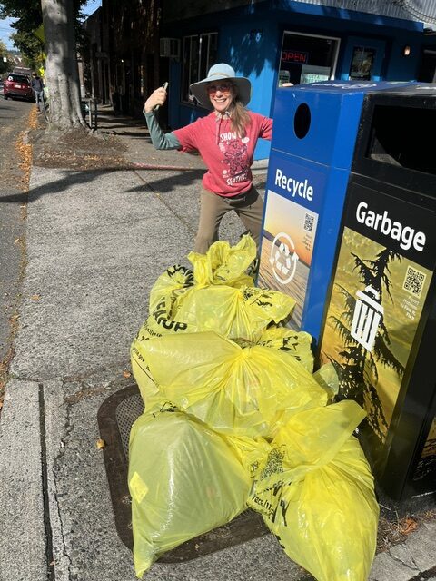 A person wearing a red shirt poses behind a large pile of yellow garbage bags next to a trash bin