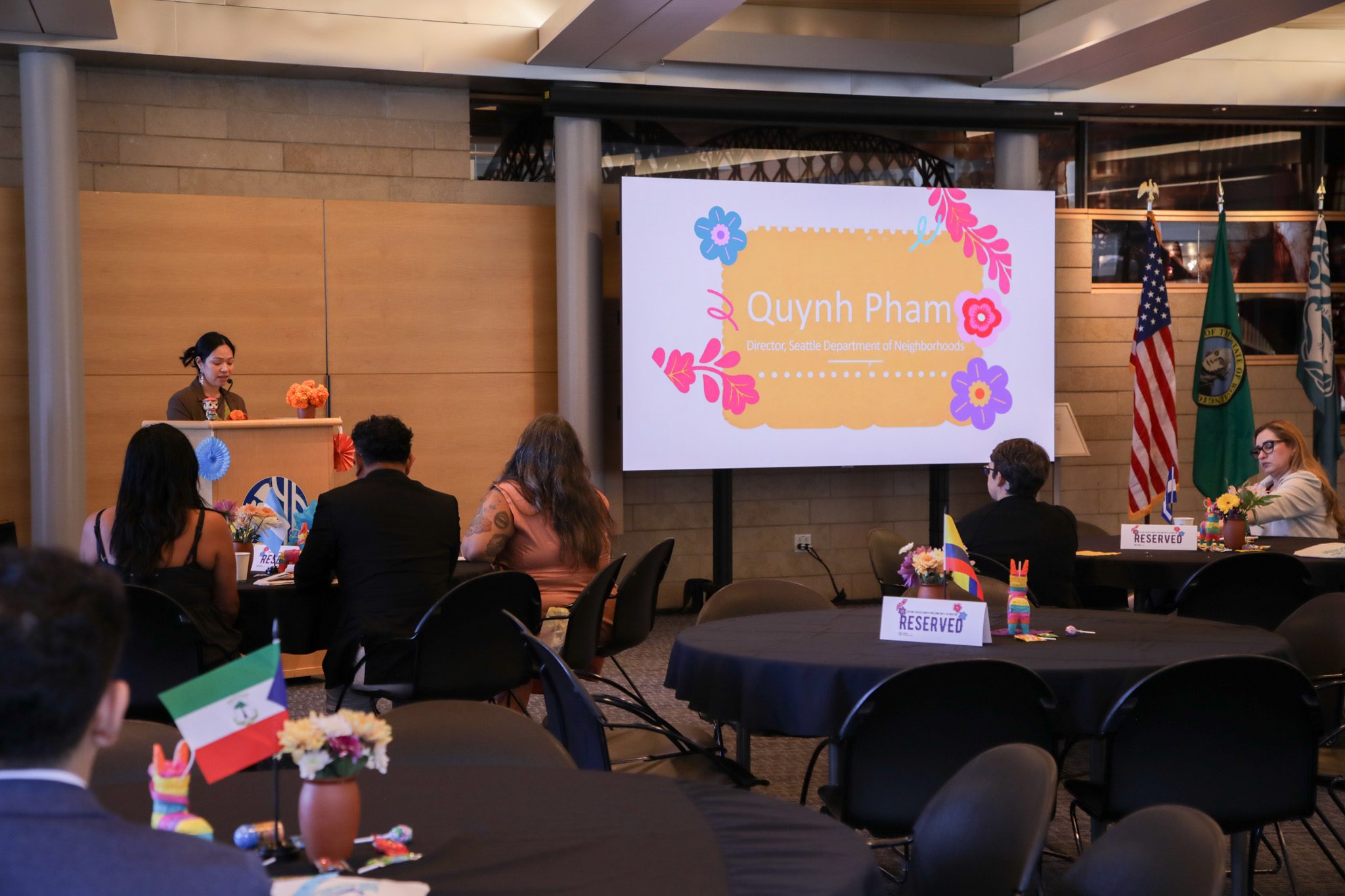 A person standing at a podium speaking to a room of people. A screen is pictured that reads "Quynh Pham, Director Seattle Department of Neighborhoods"