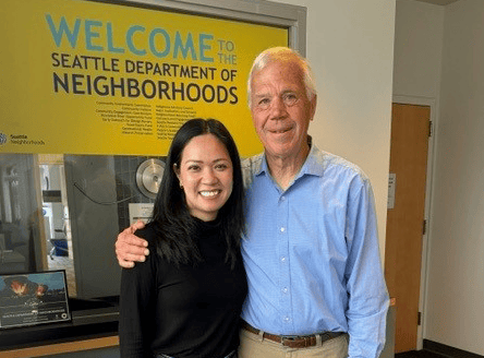 An older white man standing next to a Asian American woman in front of a sign that says "Welcome to the Seattle Department of Neighborhoods"