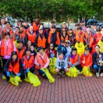 A large group of people wearing orange safety vests and holding yellow garbage sacks and garbage pickers pose in an outdoor square