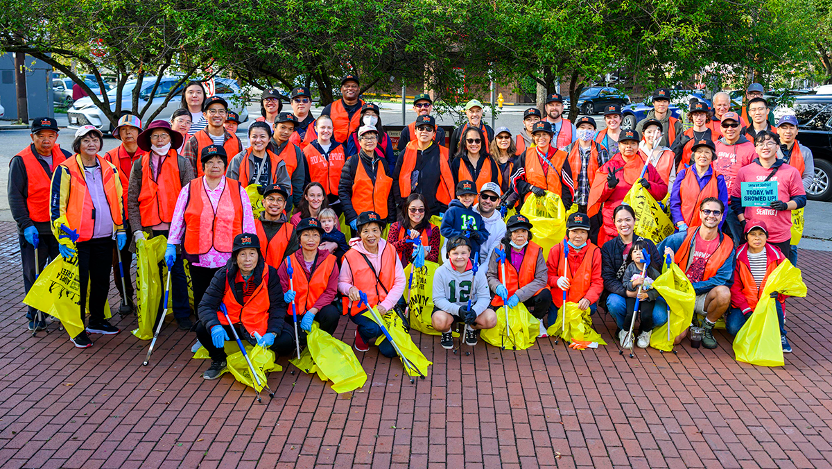 A large group of people wearing orange safety vests and holding yellow garbage sacks and garbage pickers pose in an outdoor square